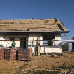 Budowa domu jednorodzinnego w trakcie realizacji: widoczne ściany z białego bloczka, drewniana konstrukcja dachu z częściowo ułożoną dachówką, rusztowania i palety z dachówką na pierwszym planie.