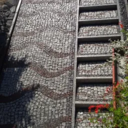 Granitowa ścieżka z mozaiką i schodami. Faliste wzory z czerwonej kostki. Detal z roślinnością. Widok z góry.