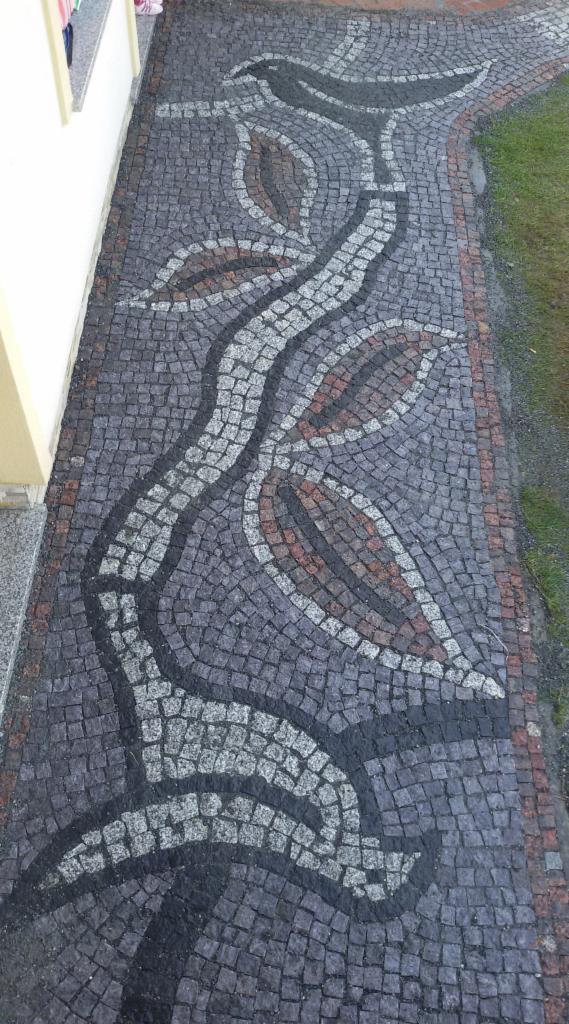 Dekoracyjna ścieżka z kostki granitowej w odcieniach szarości, czerni i brązu, ułożona we wzór pnącza z liśćmi. Widok z góry, obok budynku i trawnika.