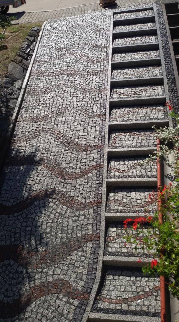 Granitowa ścieżka z mozaiką i schodami. Faliste wzory z czerwonej kostki. Detal z roślinnością. Widok z góry.