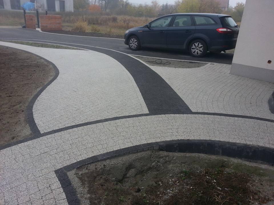 Ułożona kostka brukowa w dwóch odcieniach szarości tworząca ścieżkę i podjazd. W tle zaparkowany samochód osobowy.