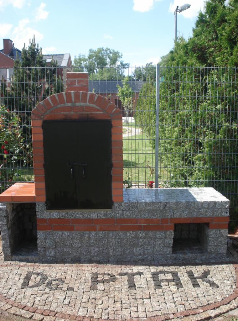 Ogród z grillem z cegły i kamienia, z napisem z kostki brukowej. Grill ogrodowy z blatem z kamienia, otoczony zielenią i ogrodzeniem.