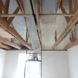 Pracownik w kombinezonie ochronnym na rusztowaniu aplikuje izolację natryskową na drewnianą konstrukcję dachu w surowym wnętrzu budynku.