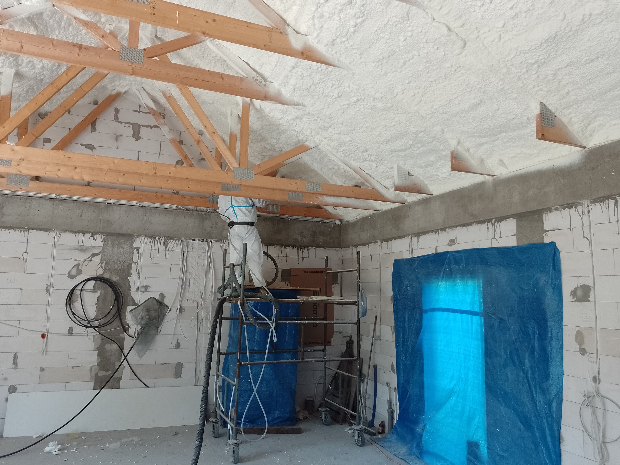 Pracownik w kombinezonie na rusztowaniu aplikuje białą izolację na poddaszu, widoczne drewniane krokwie i ściany z betonu komórkowego.