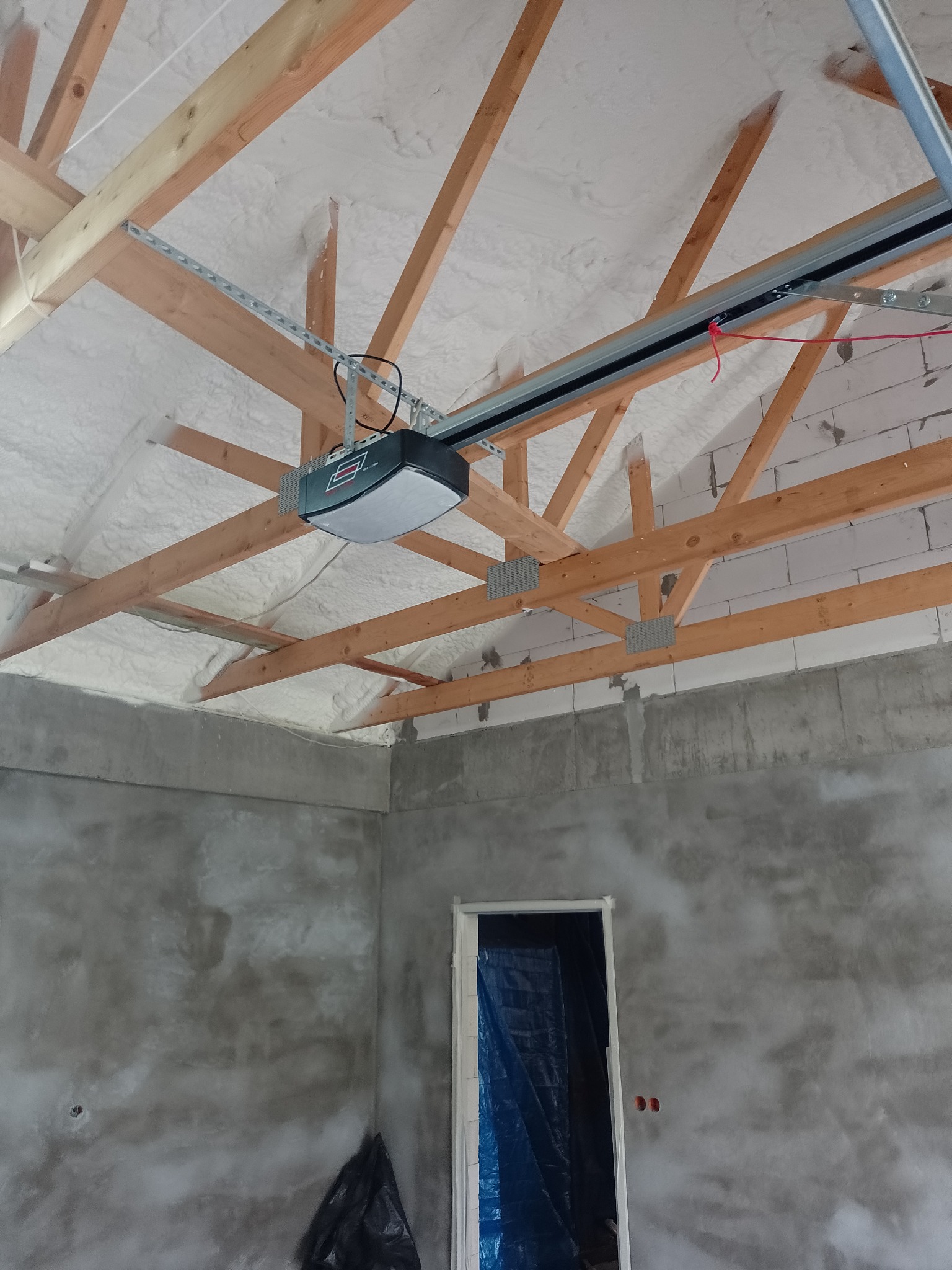 Poddasze ocieplone białą pianką natryskową, widoczne drewniane krokwie, szyny montażowe oraz automat do bramy garażowej. Ściany w stanie surowym, z widocznym tynkiem cementowo-wapiennym.