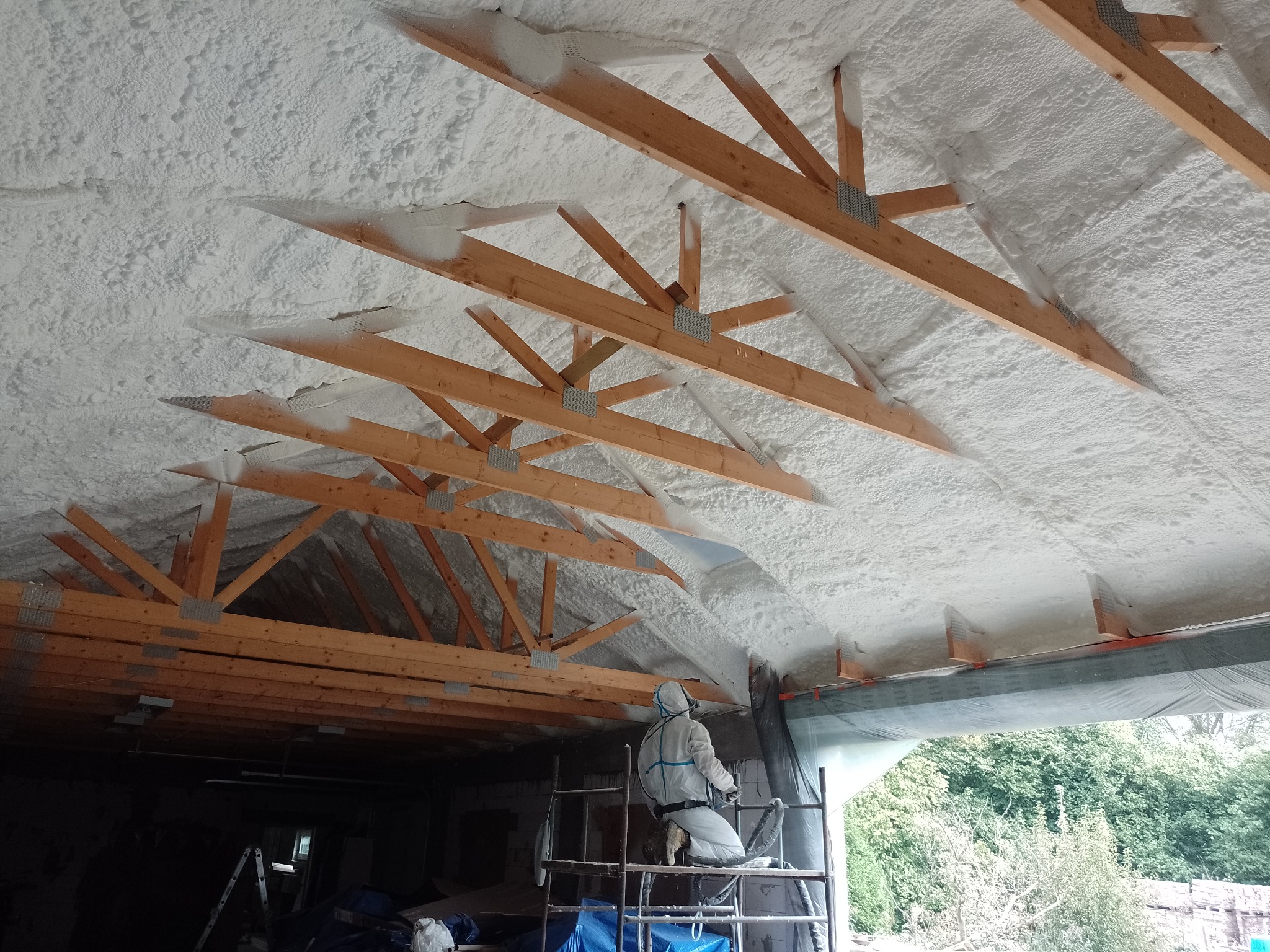 Prace ociepleniowe pianką PUR na poddaszu, widoczne drewniane elementy konstrukcji dachu oraz pracownik w kombinezonie ochronnym na rusztowaniu, aplikujący piankę.