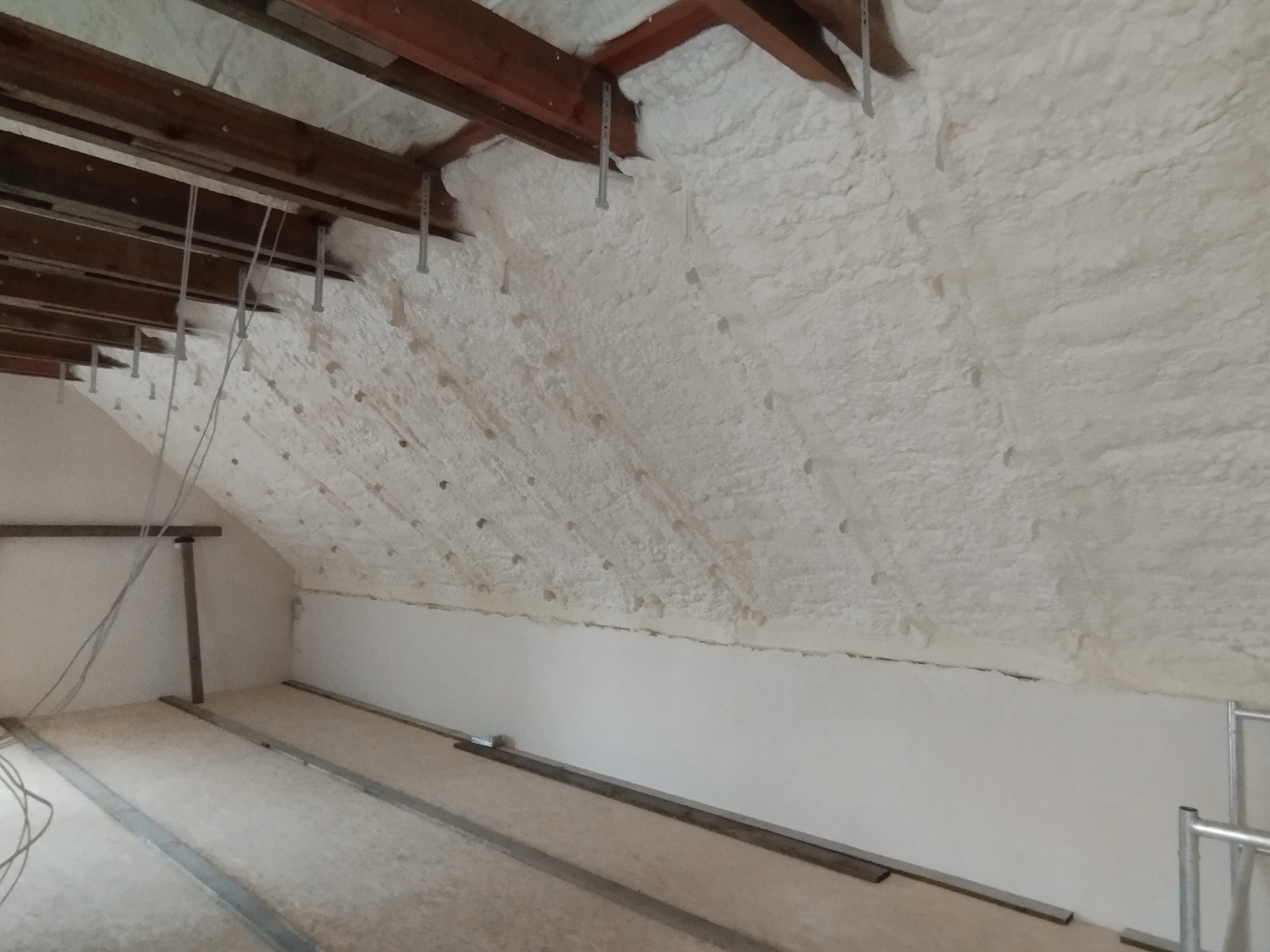 Poddasze po zaizolowaniu, widoczne drewniane elementy konstrukcyjne i metalowe wsporniki, ściany pokryte białą pianką izolacyjną.