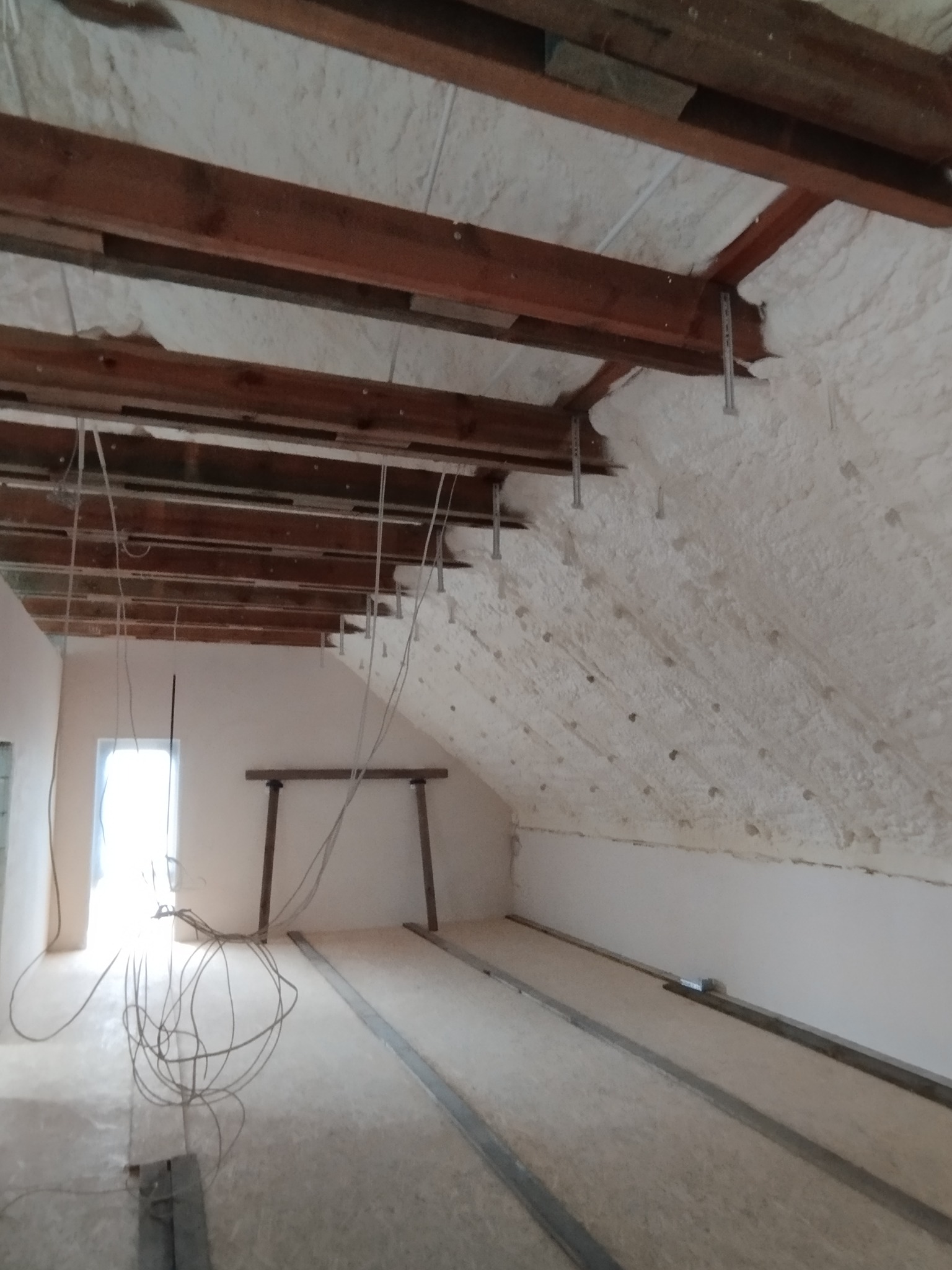 Poddasze w trakcie prac izolacyjnych, widoczne drewniane belki stropowe i biała warstwa izolacyjna na skośnej ścianie, wystające przewody elektryczne i metalowe profile podłogowe.