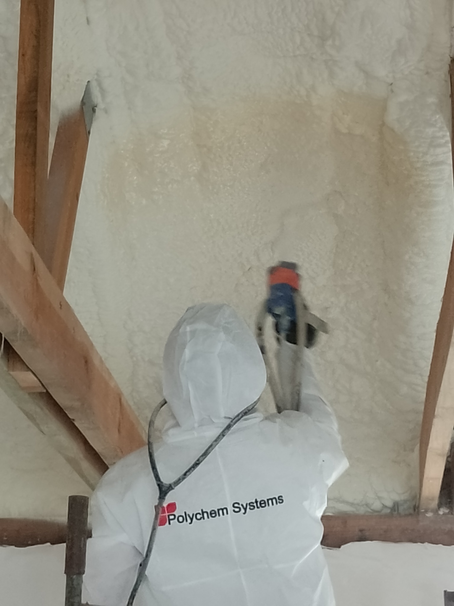 Pracownik w kombinezonie ochronnym z logo 'Polychem Systems' aplikuje piankę PUR na poddasze, widoczne drewniane elementy konstrukcyjne dachu i świeżo nałożona pianka o nieregularnej strukturze.