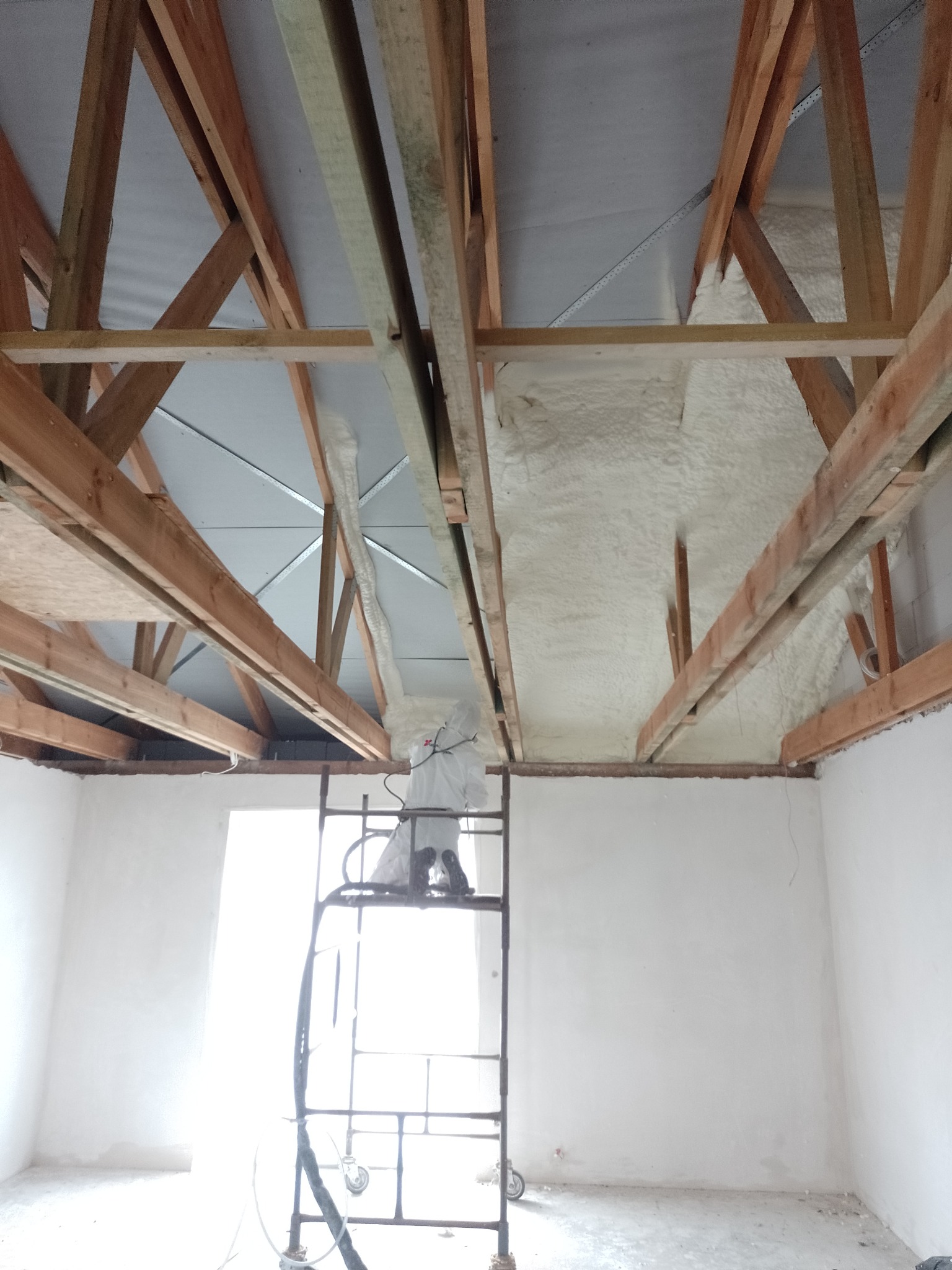 Pracownik w kombinezonie ochronnym na rusztowaniu aplikuje izolację natryskową na drewnianą konstrukcję dachu w surowym wnętrzu budynku.