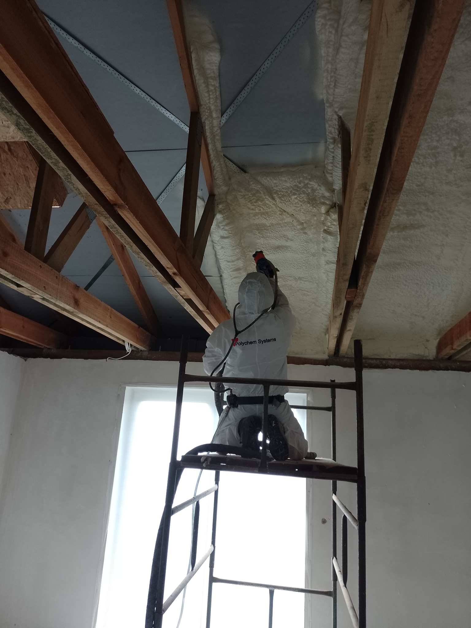 Pracownik w kombinezonie ochronnym marki Polychem Systems aplikuje izolację natryskową na poddaszu, stojąc na rusztowaniu. Widoczne drewniane elementy konstrukcji dachu i fragment okna.