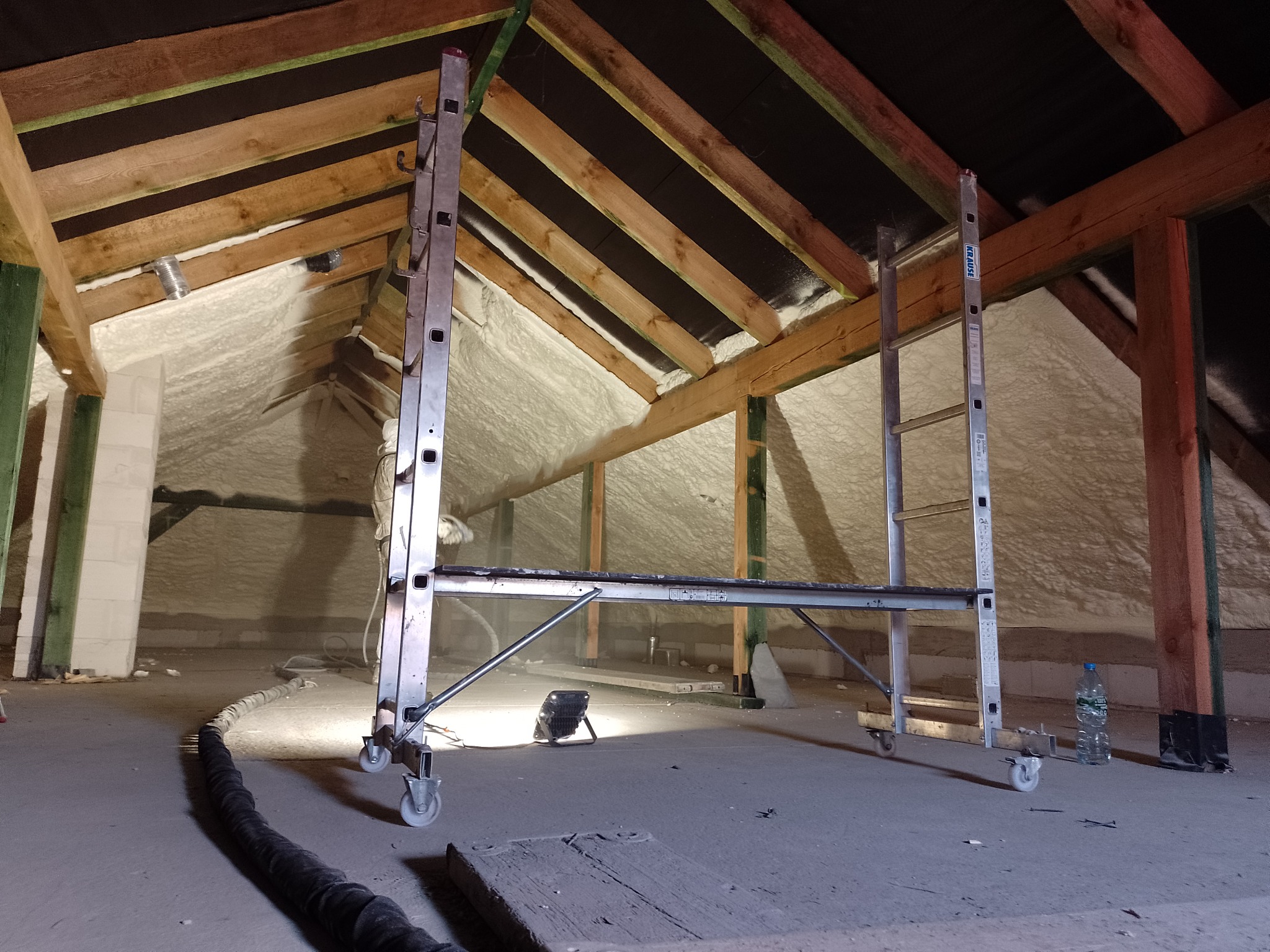 Poddasze w trakcie izolacji, pokryte pianą, widoczny aluminiowy ruszt z kółkami i wąż do natrysku piany.