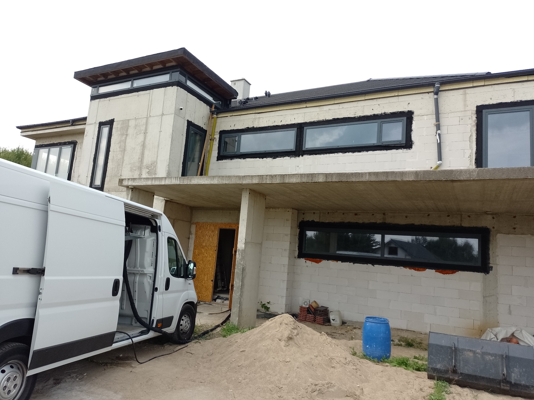 Nowoczesny dom w trakcie prac budowlanych, z widocznym samochodem dostawczym z otwartymi drzwiami i sprzętem do natrysku izolacji przy elewacji. Okna zamontowane z czarną ramą.