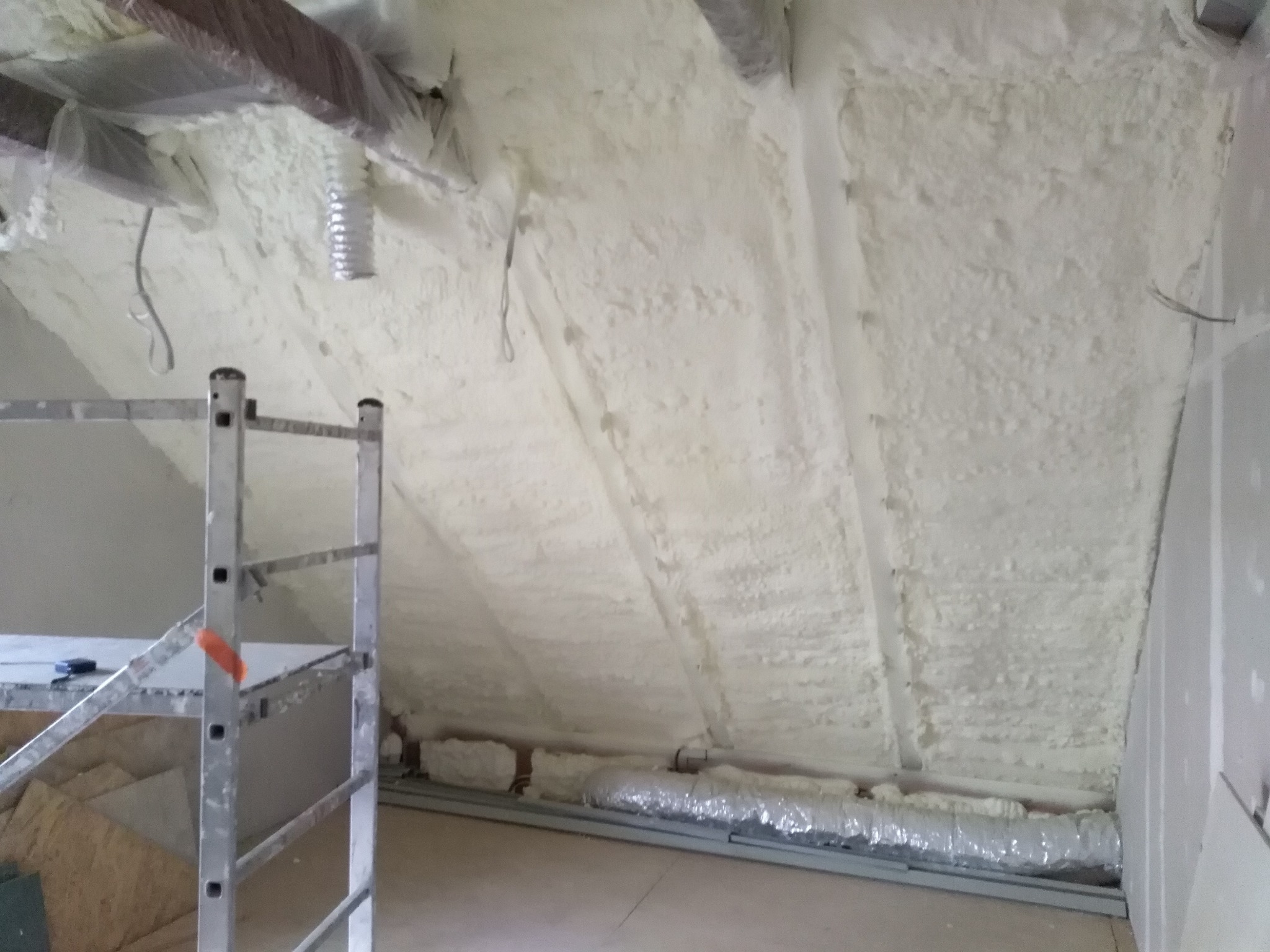 Poddasze po natrysku izolacji, widoczne drewniane belki zabezpieczone folią, aluminiowy przewód wentylacyjny oraz drabina malarska.