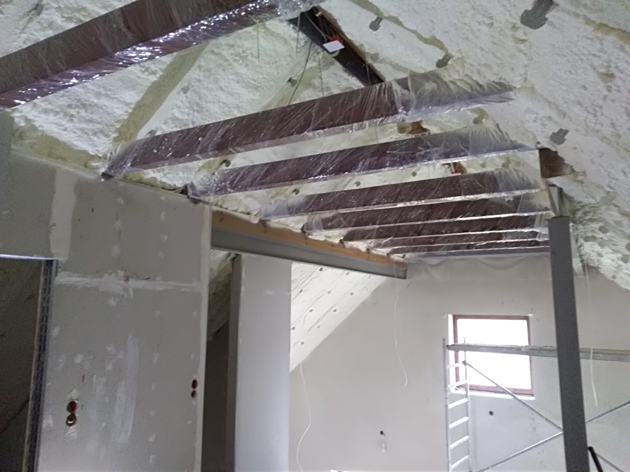 Poddasze w trakcie izolacji: drewniane belki zabezpieczone folią, pokryte warstwą pianki, widoczne fragmenty ścian w stanie surowym i rusztowanie.