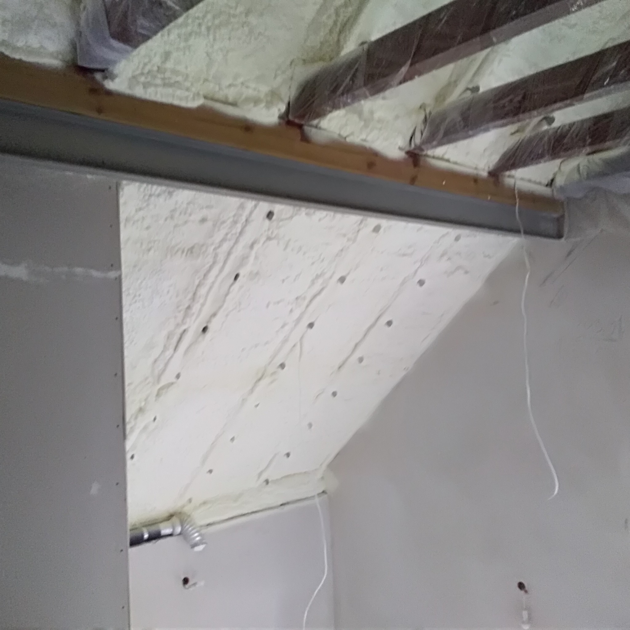 Poddasze w trakcie izolacji: widoczna pianka izolacyjna na skośnej powierzchni dachu, drewniane belki stropowe zabezpieczone folią, oraz fragment ściany z płyty gipsowo-kartonowej.