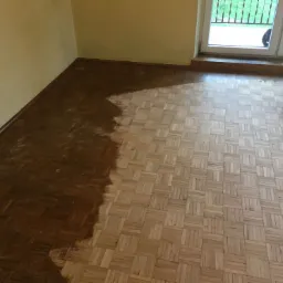 Częściowo odnowiona parkietowa podłoga w trakcie renowacji, widoczna maszyna do szlifowania, po lewej stronie ciemniejsza, odświeżona część podłogi, po prawej jaśniejsza, nieszlifowana część, żółta...