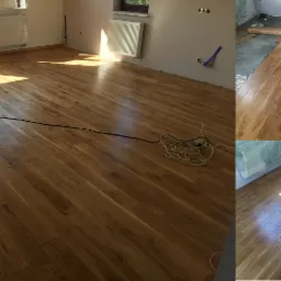 Jasne, dębowe deski podłogowe ułożone w nowym pomieszczeniu, widoczne kable i taśma malarska, w tle okno z widokiem na zieleń.