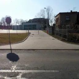 Szeroki podjazd z jasnoszarej kostki brukowej prowadzący do metalowej bramy wjazdowej, otoczony trawnikiem i ogrodzeniem z siatki, z widocznym znakiem drogowym zakazu wjazdu po prawej stronie.