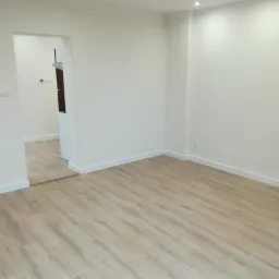 Jasne panele podłogowe w pustym, odświeżonym pokoju z białymi ścianami i listwami przypodłogowymi. Widoczne punktowe oświetlenie sufitowe i przejście do kolejnego pomieszczenia.