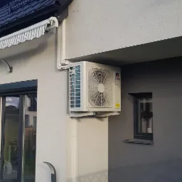 Zewnętrzna jednostka klimatyzacji marki Gree zamontowana na elewacji domu z widocznymi rurami odprowadzającymi skropliny i fragmentem instalacji elektrycznej, na tle ciemnej ściany z małym oknem.