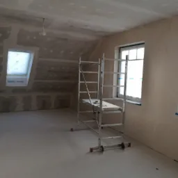 Poddasze w trakcie prac wykończeniowych, ściany i sufit pokryte płytami gipsowo-kartonowymi przed nałożeniem gładzi, widoczne rusztowanie aluminiowe, okna zabezpieczone folią malarską.