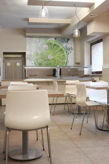 Wnętrze lokalu gastronomicznego z białymi krzesłami i stołami, oświetlone wiszącymi lampami, z dekoracyjną fototapetą przedstawiającą plaster limonki na ścianie.