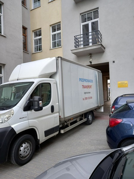 Biały bus dostawczy z napisem 'Przeprowadzki Transport Częstochowa' zaparkowany na dziedzińcu kamienicy, widoczny fragment niebieskiego i szarego samochodu.