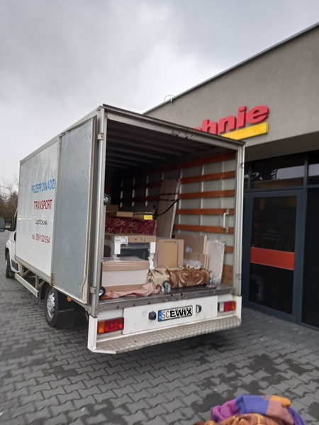 Otwarty furgon załadowany meblami i kartonami, zaparkowany na brukowanym placu przed budynkiem z szyldem sklepu, widok z tyłu pojazdu.