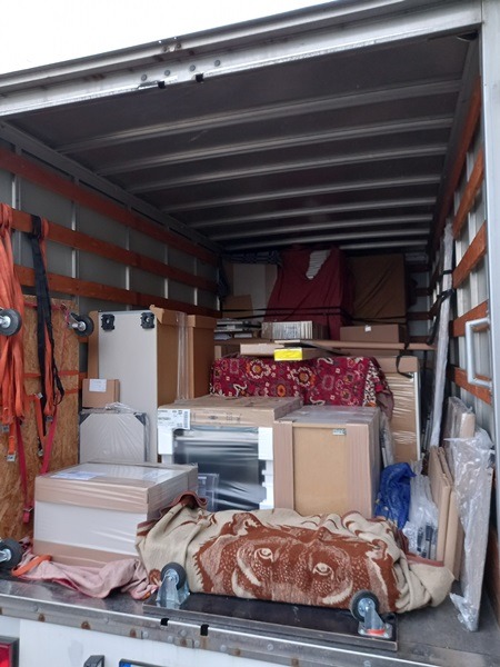 Wnętrze wypełnionej skrzyniami i meblami paki samochodu dostawczego, zabezpieczone pasami transportowymi, widoczny koc z motywem wilka na wierzchu.