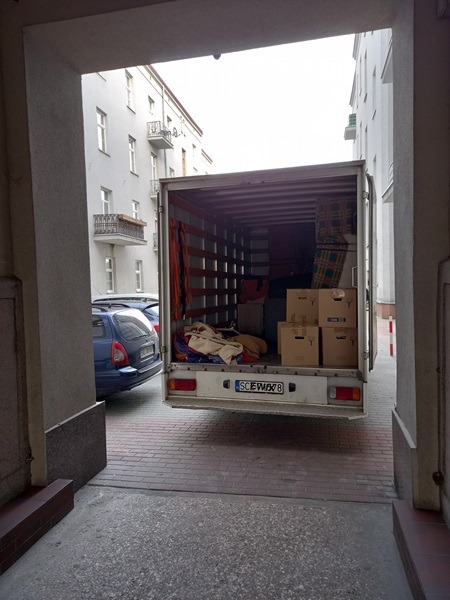 Widok z perspektywy bramy na załadowany bus przeprowadzkowy, z widocznymi kartonami i elementami wyposażenia wewnątrz, zaparkowany na brukowanym podwórku z budynkami mieszkalnymi w tle.