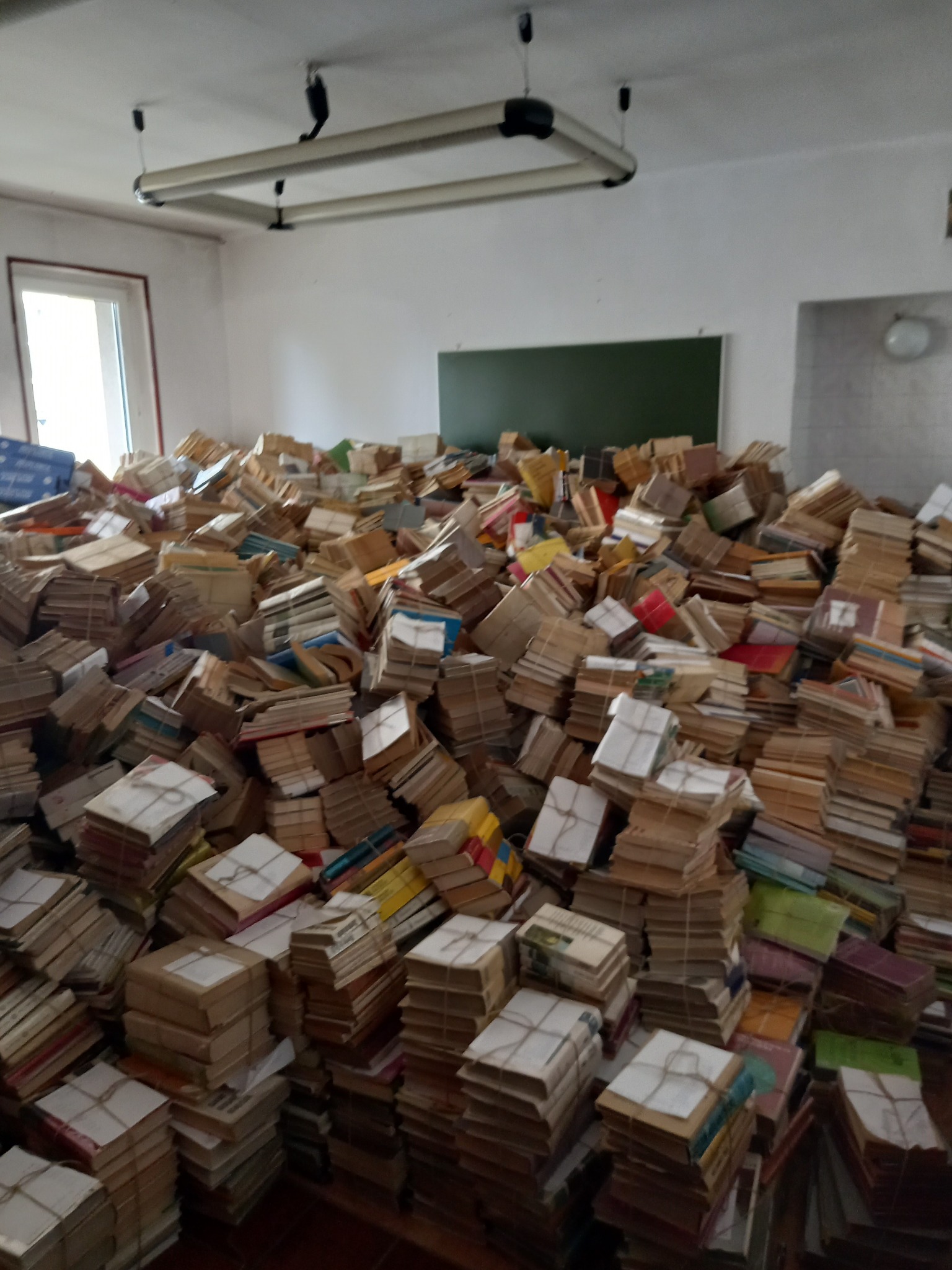 Stos książek zabezpieczonych sznurkiem, wypełniających pomieszczenie z oknem i tablicą.