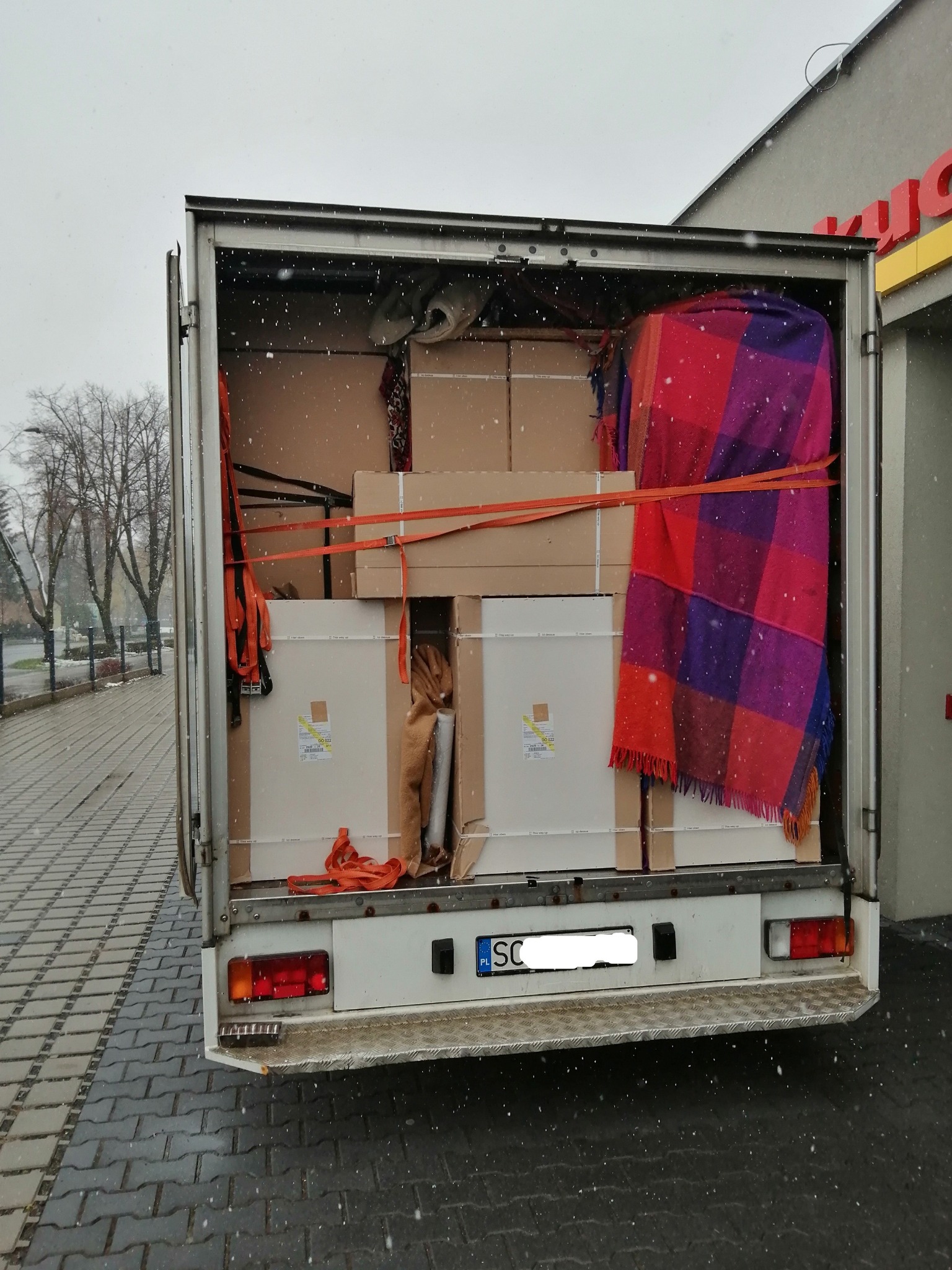 Otwarta paka busa załadowana kartonami, meblami i pledem w kratę, zabezpieczonymi pomarańczowymi pasami transportowymi, widok z tyłu pojazdu podczas opadów śniegu.