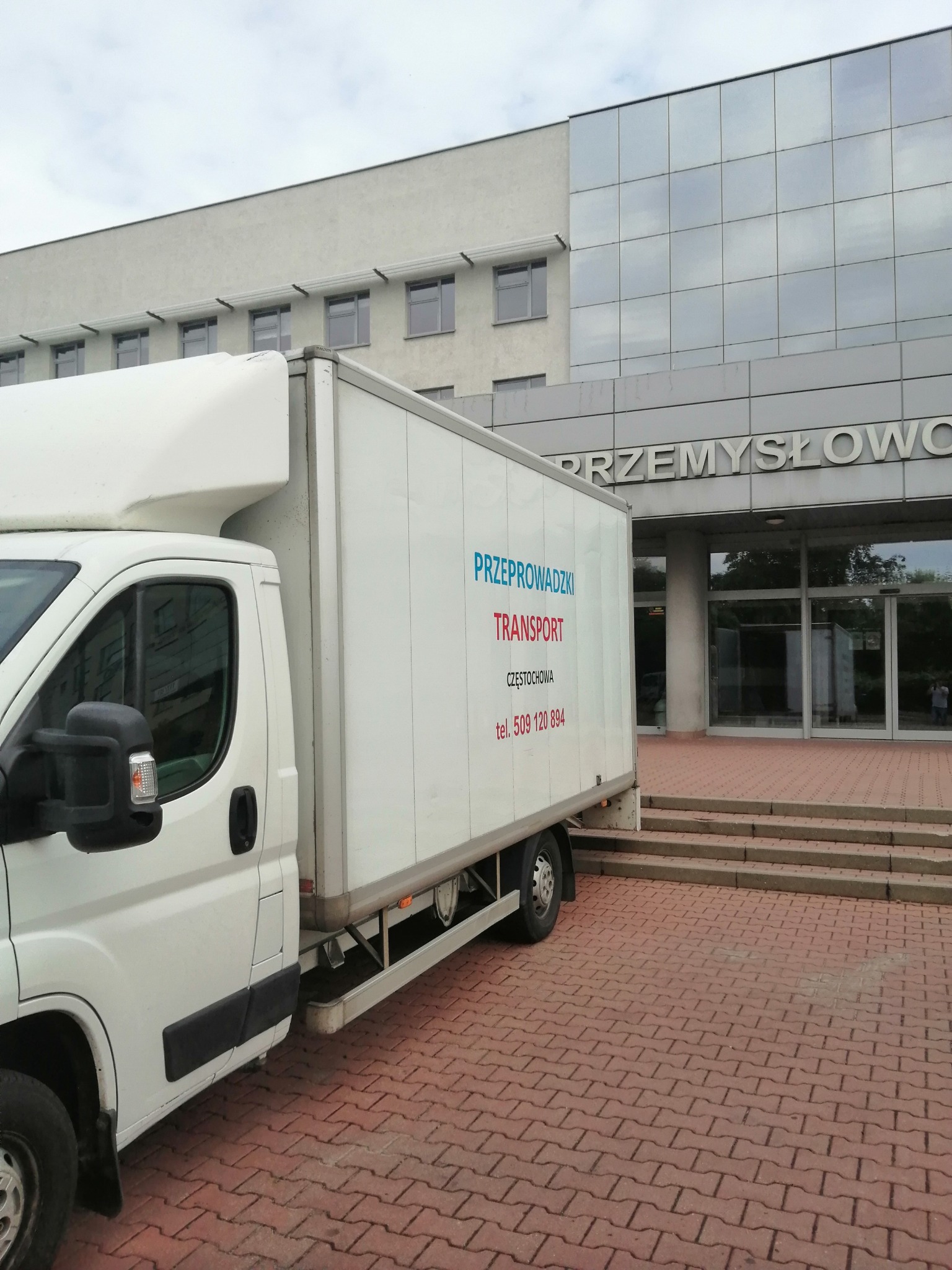 Biała furgonetka z nadrukowanym napisem 'PRZEPROWADZKI TRANSPORT Częstochowa tel. 509 120 894' zaparkowana przed budynkiem przemysłowym z przeszkloną fasadą.