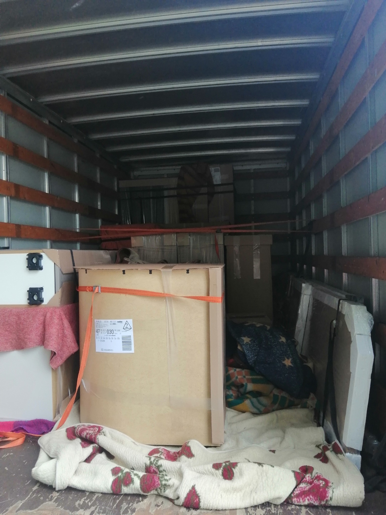 Wnętrze przestrzeni ładunkowej busa wypełnione kartonami, meblami i kocami zabezpieczonymi pasami transportowymi, widok z tyłu pojazdu.