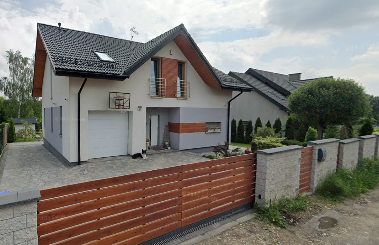 Dwukondygnacyjny dom z garażem, czarnym dachem i drewnianym akcentem. Na podjeździe leży pies, a w tle widać inne domy i zieleń. Czeladź, ul. Asnyka.