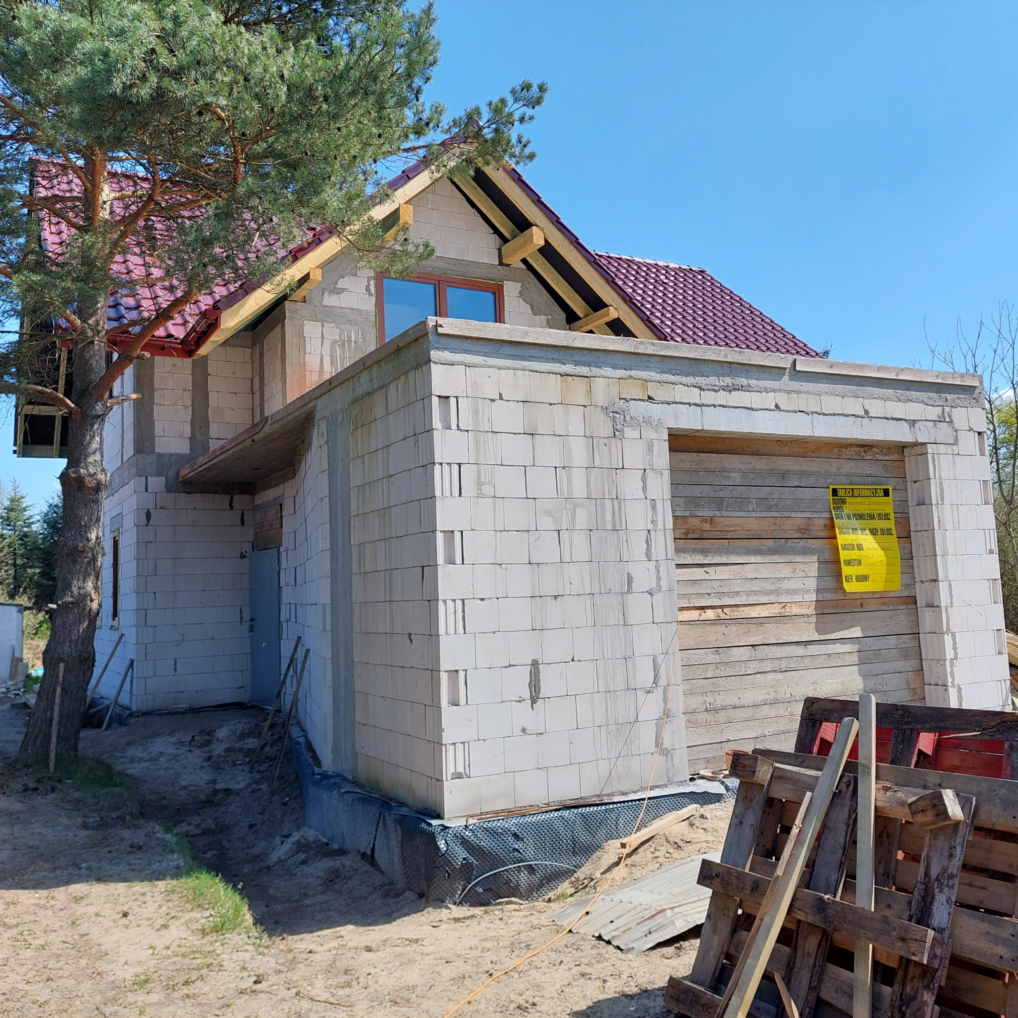 Budowa domu jednorodzinnego w trakcie realizacji: surowy mur z bloczków, czerwona dachówka, widoczna tablica informacyjna budowy, palety budowlane na pierwszym planie.