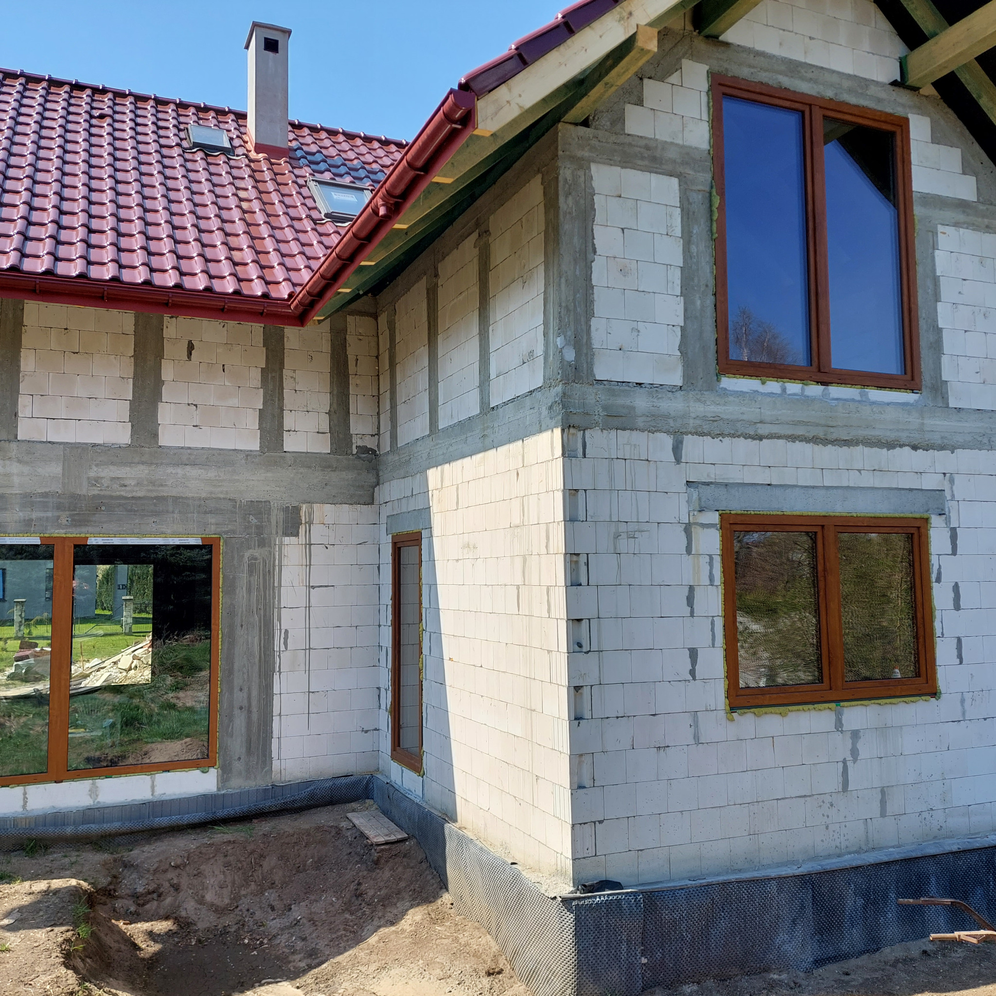 Budowa domu jednorodzinnego w stanie surowym otwartym. Widoczne bloczki, okna z brązowymi ramami, czerwona dachówka. Ujęcie z zewnątrz, w słoneczny dzień.