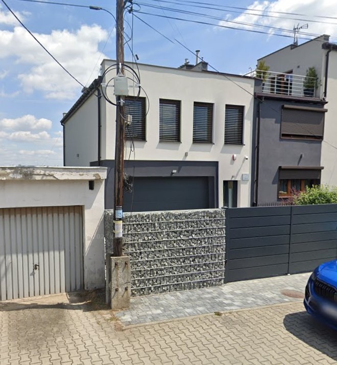 Dwukondygnacyjny dom z garażem i gabionowym ogrodzeniem na tle błękitnego nieba. Nowoczesna architektura z elementami szarości i bieli. Widok z poziomu ulicy.