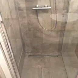 Kabina prysznicowa z odpływem liniowym w podłodze, wyłożona szarymi płytkami imitującymi kamień, widoczna bateria natryskowa i fragment umywalki przez szybę.