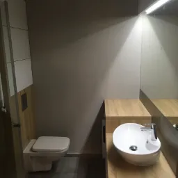 Nowoczesna łazienka z podwieszaną toaletą i umywalką nablatową na blacie imitującym drewno, oświetlona listwą LED nad lustrem.