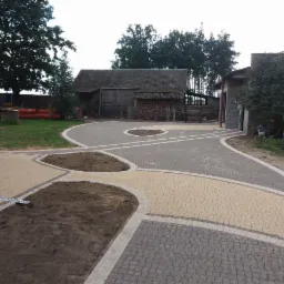 Ukończony podjazd z kostki brukowej w dwóch odcieniach szarości i beżu, z wydzielonymi miejscami na rabaty, widoczne budynki gospodarcze w tle.