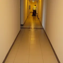 Pracownik w kamizelce odblaskowej czyści podłogę w korytarzu budynku mieszkalnego za pomocą profesjonalnego sprzętu. W tle widoczne drzwi i oświetlenie sufitowe.