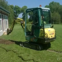 Żółto-zielona mini koparka Yanmar B15 przygotowuje wąski wykop w trawniku obok budynku z białymi oknami.