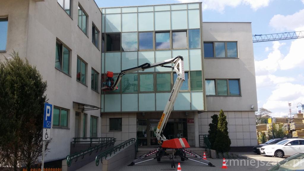 Podnośnik koszowy na tle budynku z przeszkloną fasadą, widoczne zaparkowane samochody i znak dla niepełnosprawnych.