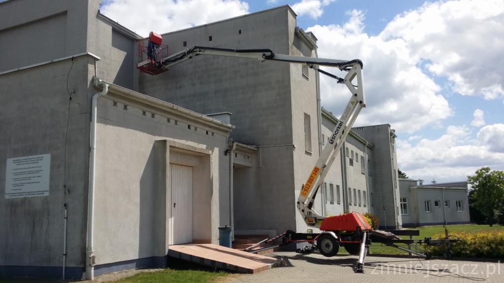 Pracownik w czerwonym koszu podnośnika teleskopowego, podczas prac na wysokości przy szarym budynku z widocznymi rynnami.