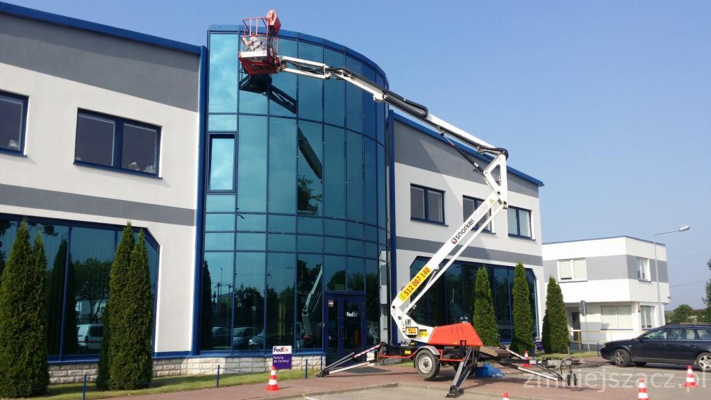 Pracownik na podnośniku koszowym, marki Snorkel, wykonuje prace na wysokości przy szklanej fasadzie nowoczesnego biurowca w słoneczny dzień.
