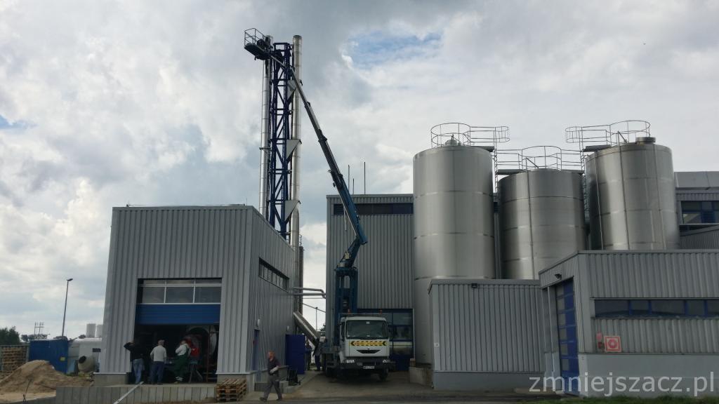 Wysięgnik podnośnika koszowego sięga do pionowej konstrukcji przemysłowej na tle zbiorników i pochmurnego nieba. Grupa osób przy budynku. Widoczny samochód ciężarowy.