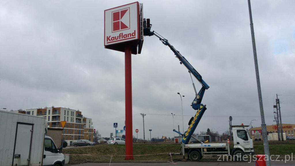 Montaż lub konserwacja szyldu reklamowego Kaufland za pomocą podnośnika koszowego na tle pochmurnego nieba i zabudowań miejskich.