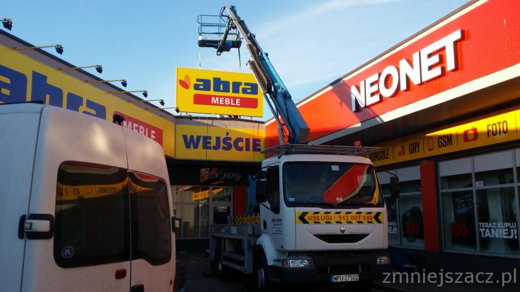 Podnośnik koszowy przy montażu lub demontażu reklamy zewnętrznej na budynku sklepu, widoczne logotypy 'Abra Meble' i 'Neonet'.